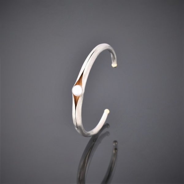 Upright picture of folding anticlastic matt silver cuff holding a freshwater pearl. Gold detailing on tips